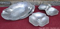 Photo showing all five vintage aluminum serving pieces by Trade Continental arranged on a flat surface, highlighting the varying shapes and sizes with floral and leaf embossed hand-hammered texture.