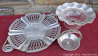 Photo of three aluminum serving pieces: a hand-wrought tray with decorative cutouts and spiral handles, a bowl with a glass bottom and fluted rim, and a small nut dish with a floral handle.