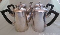 Four vintage silver-plated coffee serving pots with black plastic handles grouped together on light background menunjukkan wear including tarnish and scratches.