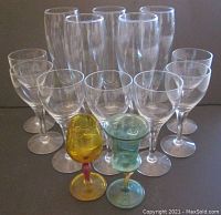 Front view of 14 champagne and cordial glasses arranged on surface, showing mostly clear glassware with two colored cordial glasses in front.