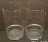 Pair of large, clear cylindrical glass vases with straight sides shown side by side on a dark surface.