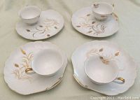 Top-down view showing the four sets of white snack plates with matching tea cups with hand painted golden wheat design and gold accents.