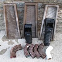 Three brown plastic splash blocks standing vertically at back, two flex grate adjustable downspout filters with attached flexible ducts in front, six elbows including five brown and one white aluminum elbow laid out on ground.