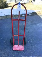 Front view of the red two-wheel dolly standing upright on asphalt with snow background, showing the curved handle and pneumatic tires.