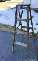 Full view of the 3-step wooden stepladder standing outdoors on pavement, showing its structure and condition including missing paint tray holder.