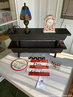 View of two dark wooden benches stacked, ceramic floral clock, stained glass lamp, patriotic sign, cross stitch, circular glass candle plate on white surface.