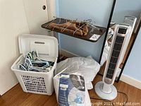Photo shows Lasko tower fan, bed/chair tray table, white laundry basket filled with assorted hangers, two mattress pads, and a full-length mirror.