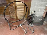 Tubular metal log holder, fireplace accessories including andirons, tools on stand, bellows, and gloves arranged on floor against background wall.