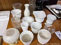 Photo of assorted milk glass vases including textured and smooth styles, one glass lidded jar, and one black ceramic vase, arranged on wood surface