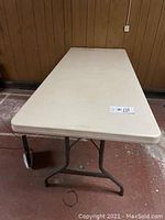 Full view of rectangular beige plastic table with metal legs in folded position against wood panel wall.