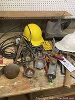 Wide view of lot showing three smelting ladles, two hard hats, various hand tools, and vintage electric light on wooden surface.