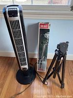 Lasko tower fan next to Solidex tripod box and tripod on wooden floor near window