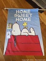 Photo of the faded fabric flag with Snoopy lying on top of a red doghouse with Woodstock, with 'HOME SWEET HOME' text across the top.