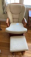 Photo showing the full tan upholstered glider chair with wooden arms and matching hassock below, positioned on hardwood floor near window.