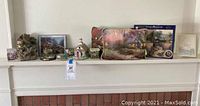 Wide shot of mantel displaying various Thomas Kinkade themed items including puzzle boxes, picture frames, and decorative houses.