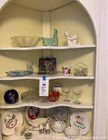 Four shelves displaying a variety of Depression glass, souvenirs, pottery, and china including clear and colored glass dishes, floral and map-themed plates, planters, and other assorted decorative items.