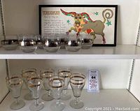Shelf display showing eight clear roly-poly glasses with silver zodiac bands on top shelf, and seven goblets with gold bands below alongside Taurus traits plaque.
