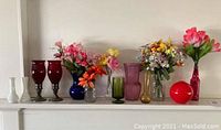 Wide shot of colorful glass vases and ruby glass candleholders on a white shelf with artificial spring flowers.
