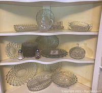 Shelf display with more than twenty five pieces of clear Fostoria Americana glassware including various plates, serving dishes, salt and pepper shakers, and candy dish with lid.