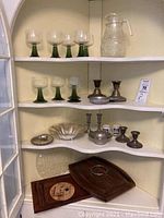 Cabinet shelf displaying nine green stemmed glasses, clear embossed glass pitcher, three pairs of candlesticks including sterling silver, and wooden serving trays.