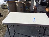 White rectangular plastic folding table with metal folding legs on asphalt surface, showing size and general condition.