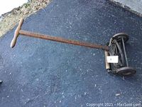 Full view of the vintage push reel lawn mower placed on pavement showing metal reel, three metal wheels, and wooden handle with T-shaped grip.