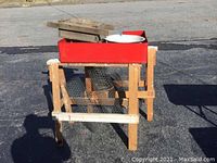 Pair of wooden saw horses supporting a red metal tray with a white enamel bowl on top, with pieces of chicken wire visible underneath.