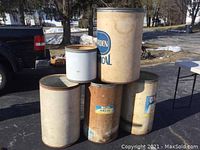 Five cardboard industrial drums of various sizes stacked outdoors in sunlight. Drums show signs of wear and staining.