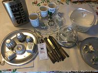Overview of the entire lot showing mugs, metal molds, serving platter, CorningWare casserole dish, cheese dome, knives, metal container and tongs.
