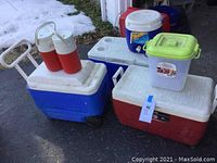 Photo showing four beverage coolers and containers including blue wheeled cooler, red and white cooler with cup holders, white and red cooler, and green topped container.