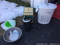 Full lot view showing 30-cup electric coffee percolator, large stainless steel strainer, green plastic bowl, white laundry hamper with lid, and a small cardboard box, all placed outdoors on pavement.