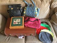 Photo showing vintage Taperlite hard sided suitcase, black handbag, blue quilted Vera Bradley handbag, Tennessee Wood Crafters wooden storage box with quilted cover, and multiple smaller bags in pink and other colors arranged on a beige couch.