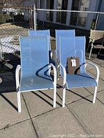 Two blue fabric deck chairs with white painted metal frames shown side by side outdoors on concrete, sunlight casting shadows. Chairs have curved armrests and slightly reclined backs.