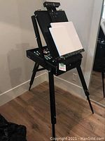 Black collapsible tripod artist easel set up with storage box and blank canvases