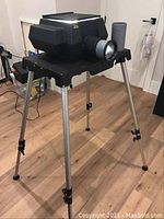 Front-side view of the black Artograph Super AG 100 projector mounted on adjustable silver tripod stand, showing projecting lens and second lens attachment.
