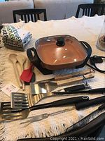 Starfit electric frying pan with glass lid, wooden spoons, red silicone spatula, BBQ tools, and kitchen tea towels on a table.