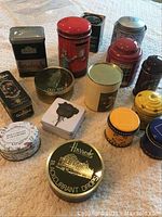 Photo showing 16 assorted collector tins of various shapes, sizes, and colors on a patterned cloth surface.