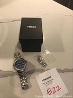 Full lot showing the Timex watch, its original black box, extra metal links in a small plastic bag, and MaxSold card.