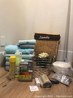 Photo showing wicker laundry hamper with fabric lid marked 'Laundry', stacked towels in turquoise and gray, assorted cleaning products, foam soap bar, small decorative flower piece, metal frame, lint roller, and roll of fabric or paper.