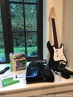 Full view showing Xbox 360 console, Kinect sensor bar, black guitar controller, stacked games and manuals on a white surface by window.