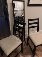 Two chairs with dark brown or black wooden ladder backs and cream cushioned seats placed near a wall mirror with a black frame.