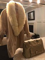 Cream women's coat with a thick, fluffy white faux fur collar hanging on a hanger with the beige handbag placed in front on a table under warm lighting.