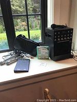 Black karaoke main unit on windowsill with one wireless microphone on top, RCA cables and carry bag to the side.