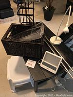Wide view of miscellaneous household items including woven basket, black side table, white stepping stool, desk lamp, and curtain rods.