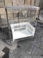 Metal wire shelving unit with three wire shelves and white plastic bin on bottom shelf, photographed outside on stone tile surface.
