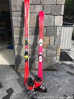 Photo of two pairs of skis (Atomic 180 cm red and Head 150 cm red/pink) standing upright with ski boots in front.