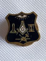 Close-up photo of the antique Masonic pin showing the shield shape, black enamel, and gold-tone Masonic symbols including the all-seeing eye, compass and square with 'G', plumb, hourglass, and trowel.