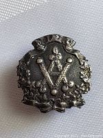 Close-up photo of the antique Masonic silver pin showing front detail with square and compasses and 'G' symbol surrounded by floral wreath.