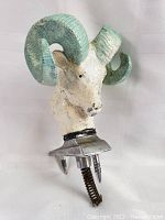 Three images showing a metal vintage RAM hood ornament from multiple angles. The RAM has turquoise painted large curved horns and a white face with some wear and paint chipping. Mounted on a metal base with mounting screws and spring attachment.