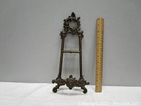 Full view of brass tabletop easel next to ruler showing its measured height around 12 inches. The easel shows an ornate design with scrolls and floral motifs, with one leg missing making it incomplete.